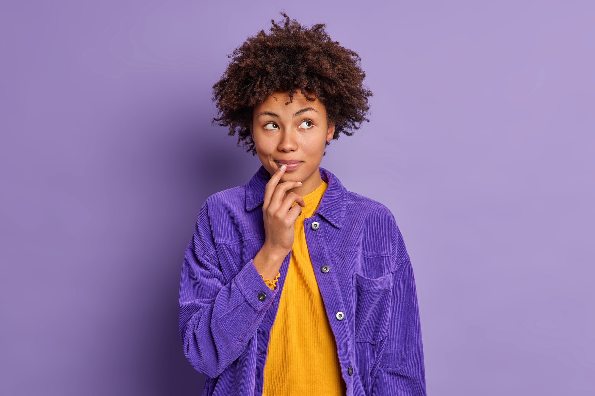 studio-shot-dreamy-dark-skinned-young-woman-keeps-hands-near-mouth-looks-thoughtfully-aside-dressed-stylish-violet-jacket